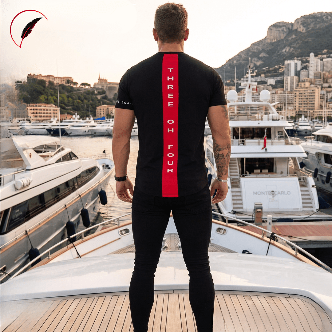 A man stands on the deck of a yacht in a marina, viewed from behind, wearing a black t-shirt with a bold red vertical stripe on the back featuring the text 'THREE OH FOUR'. The background shows luxury yachts and a coastal cityscape.