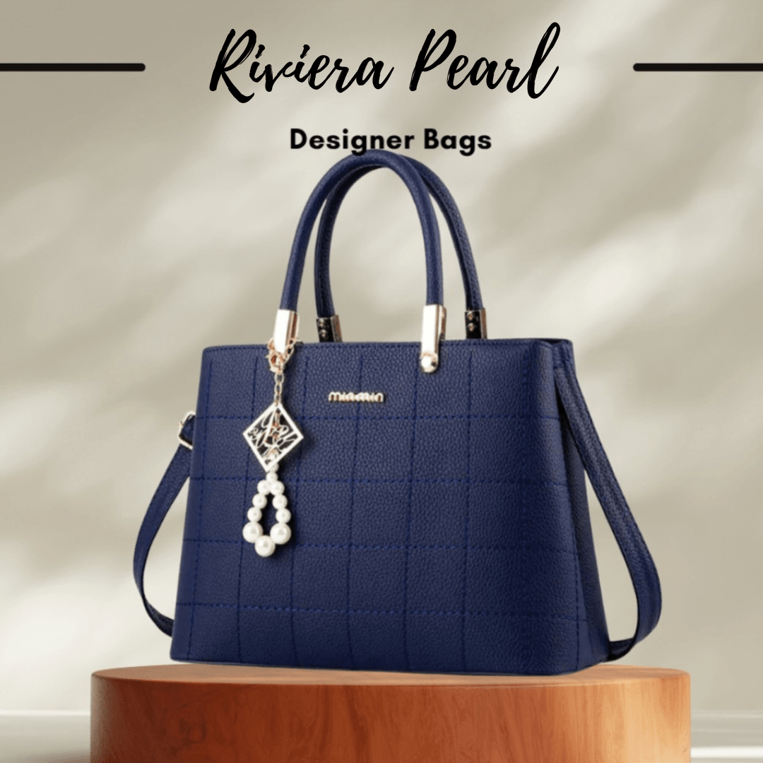 A navy blue textured handbag with double top handles, a shoulder strap, and a dangling pearl charm, displayed on a wooden pedestal with the text 'Riviera Pearl Designer Bags' above it.