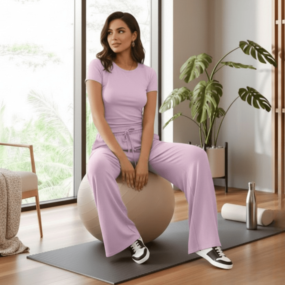 A woman sits on a beige exercise ball in a bright room, wearing a matching light purple/lavender athletic set consisting of a short-sleeve top and wide-leg pants. She is positioned on a dark gray yoga mat, with a plant, window, and home gym accessories visible in the background.