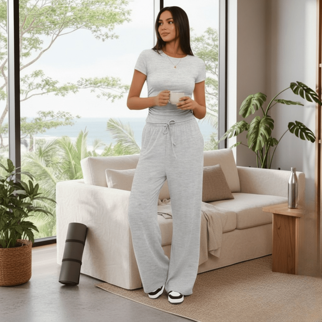 A woman stands in a bright living room wearing a light gray short-sleeve t-shirt and matching wide-leg sweatpants, holding two white mugs. The room features a beige sofa, potted plants, and a large window overlooking a tropical beach.