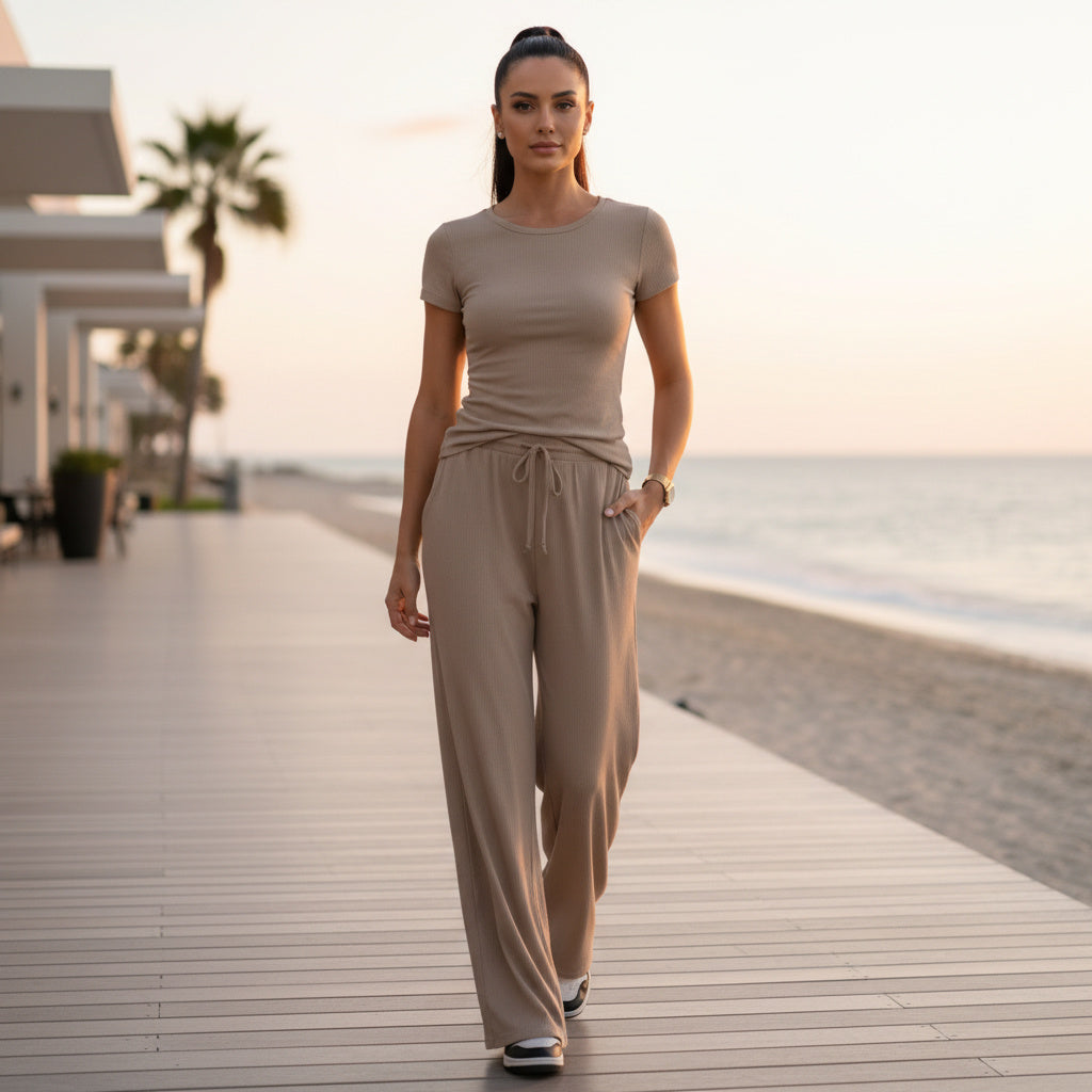 A woman walks on a wooden boardwalk beside the ocean at sunset, wearing a matching beige short-sleeve top and drawstring wide-leg pants.