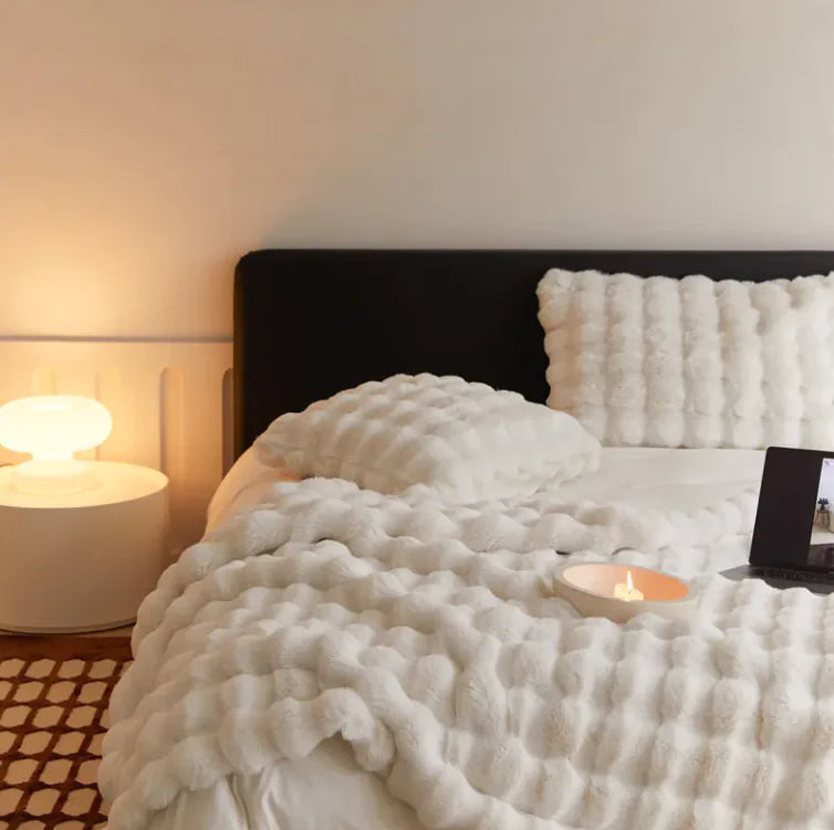 Bed with white textured blanket and pillows, nightstand with lamp, and candle on a neutral background