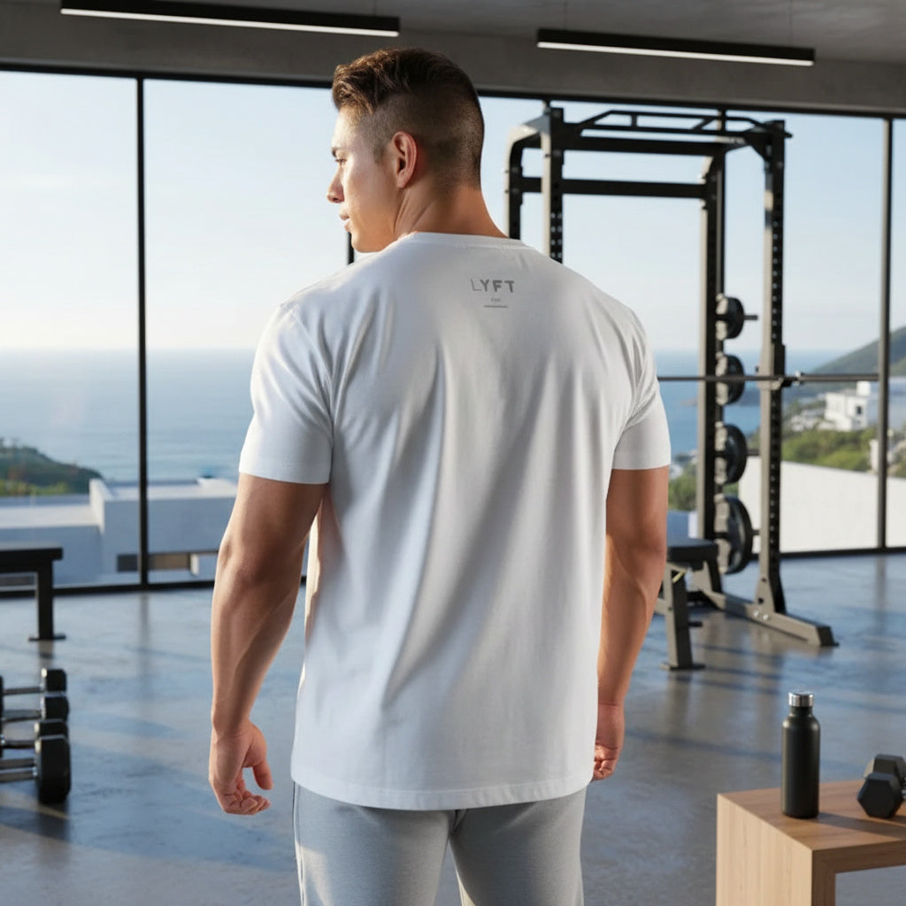A man in a gym wearing a white short-sleeve t-shirt with a small gray 'LYFT' logo on the upper back, shown from behind. The gym has large windows overlooking the ocean, weight machines, and a wooden table with a water bottle.