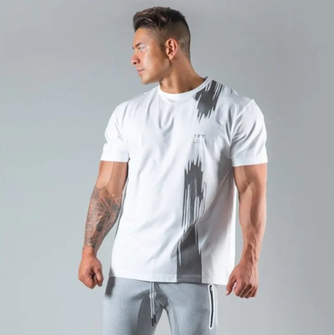 A muscular man wearing a white short-sleeve athletic t-shirt with a gray vertical brushstroke pattern on the front, paired with gray sweatpants, set against a plain gray studio background.