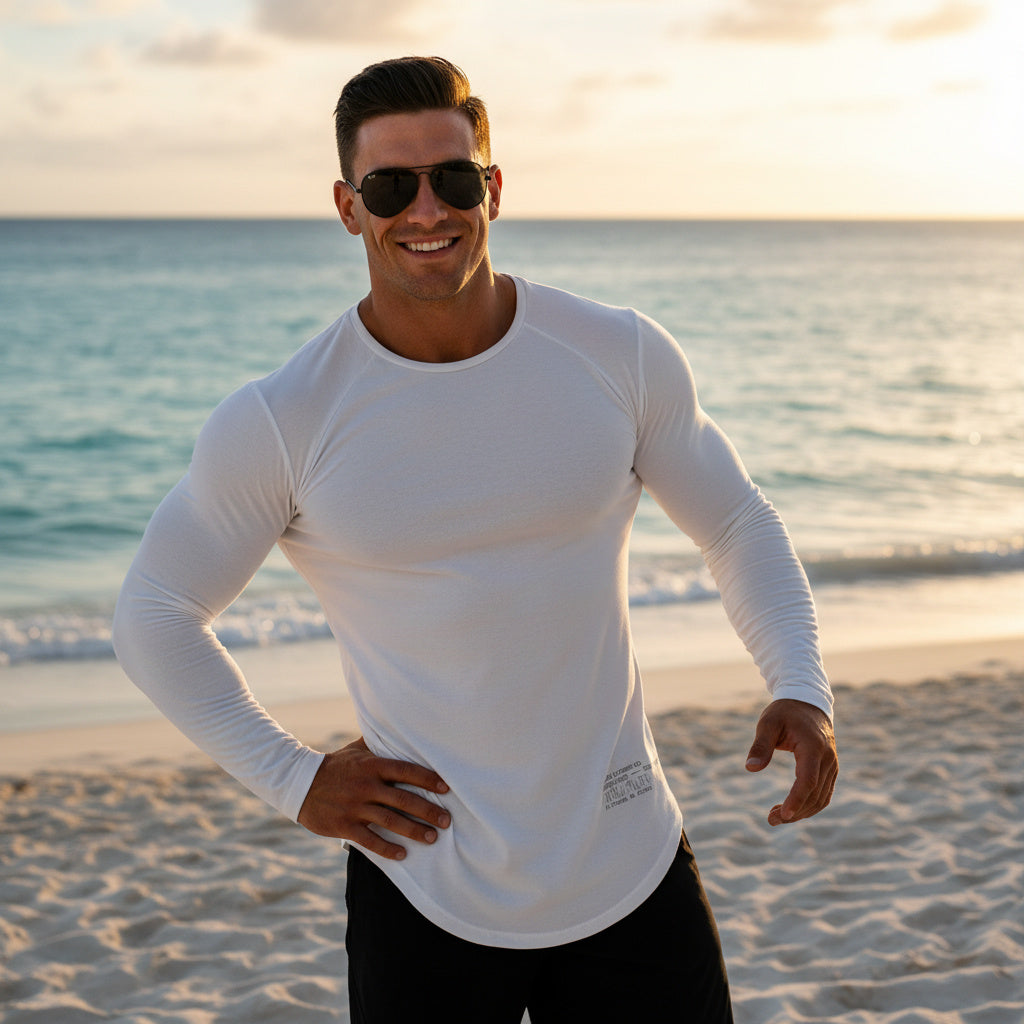 Man wearing a white long-sleeve shirt and sunglasses on a beach at sunset.