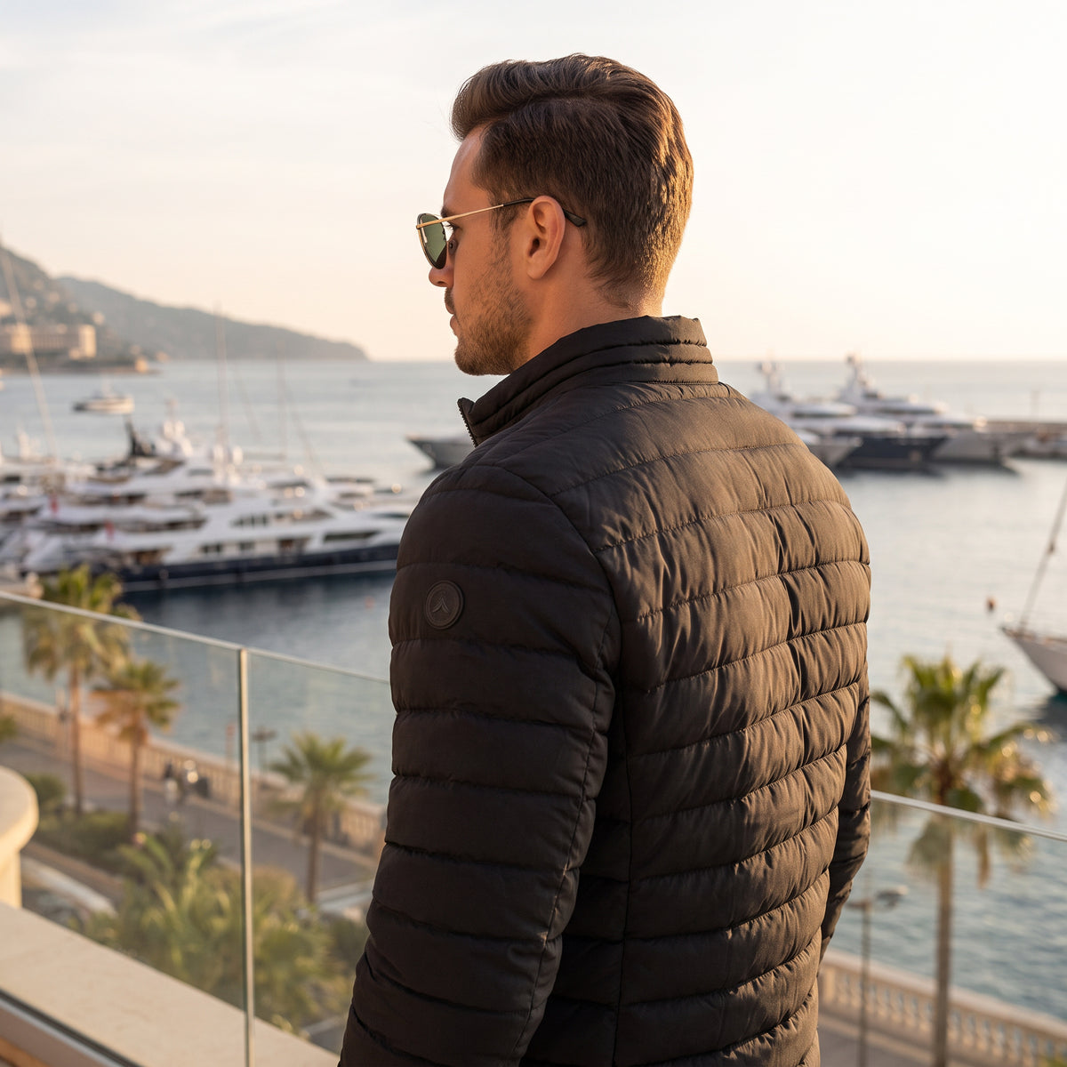 A man wearing a black quilted puffer jacket and sunglasses stands on a balcony overlooking a marina with yachts and water at sunset.
