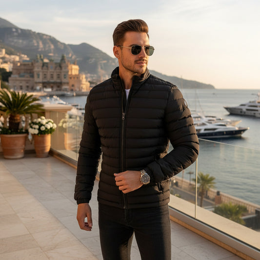 A man wearing a black quilted puffer jacket and sunglasses stands on a balcony overlooking a coastal city and marina at sunset.