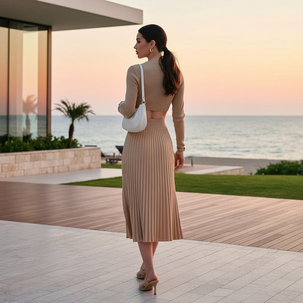 Woman in a beige dress standing on a wooden deck overlooking the ocean at sunset.