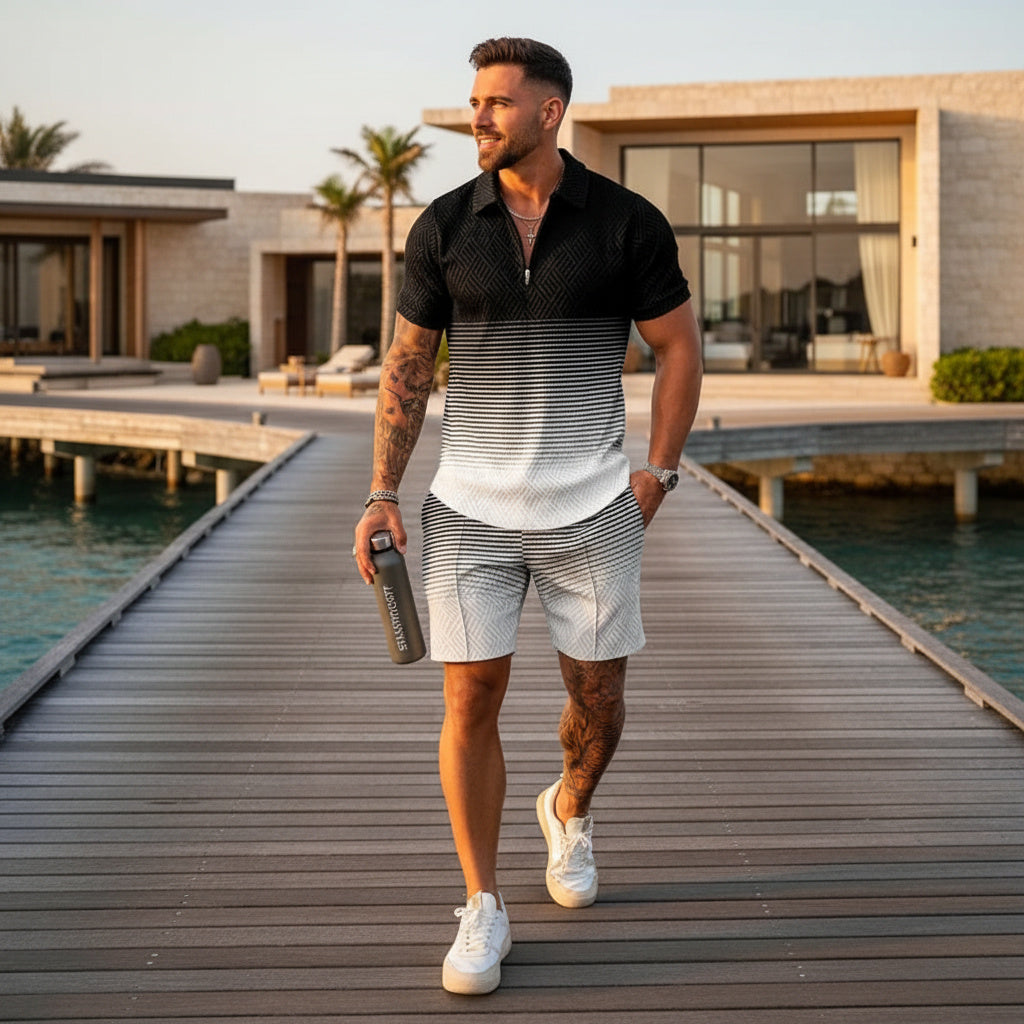 A man walks on a wooden dock by the water wearing a black-to-white gradient short-sleeve polo shirt, white patterned shorts, and white sneakers, with a modern villa in the background.