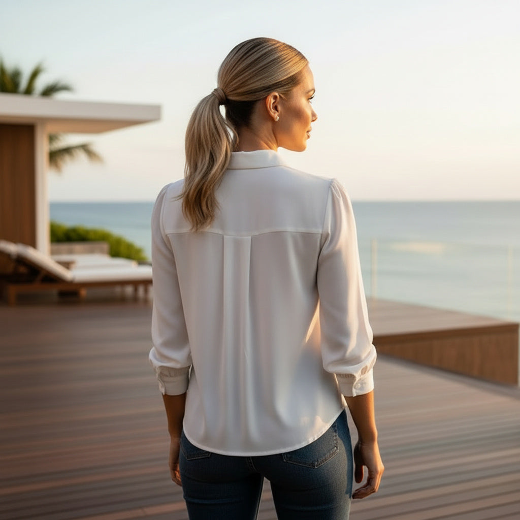 A woman with blonde hair in a ponytail stands on a wooden deck overlooking the ocean, wearing a white long-sleeve blouse and dark blue jeans.