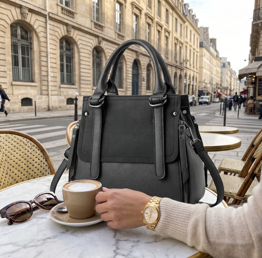 A black leather handbag sits on a Parisian cafe table with a person holding a coffee cup, with the street and buildings in the background.