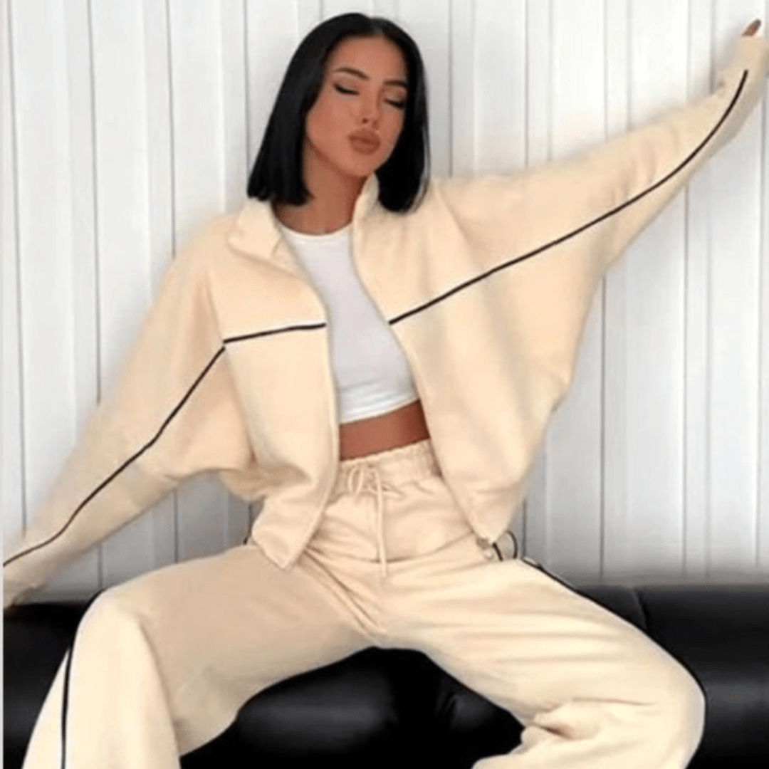 A woman wearing a cream-colored two-piece tracksuit with black piping details, consisting of a cropped zip-up jacket and matching sweatpants, posing on a black leather couch against a white paneled wall.