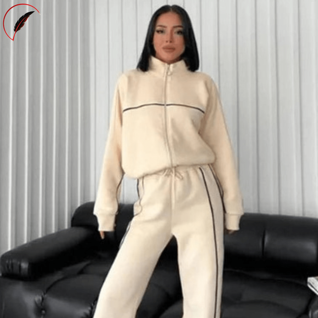 A woman modeling a cream-colored two-piece tracksuit with thin black stripe details, standing in front of a white pleated curtain and a black leather couch.