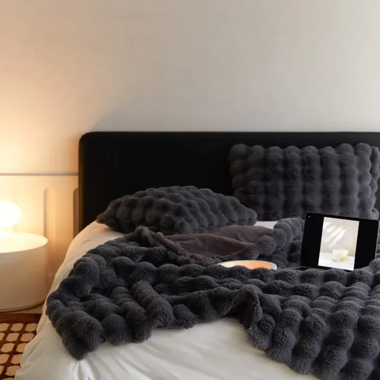 Dark gray textured blanket draped over a bed with a nightstand and lamp in the background.