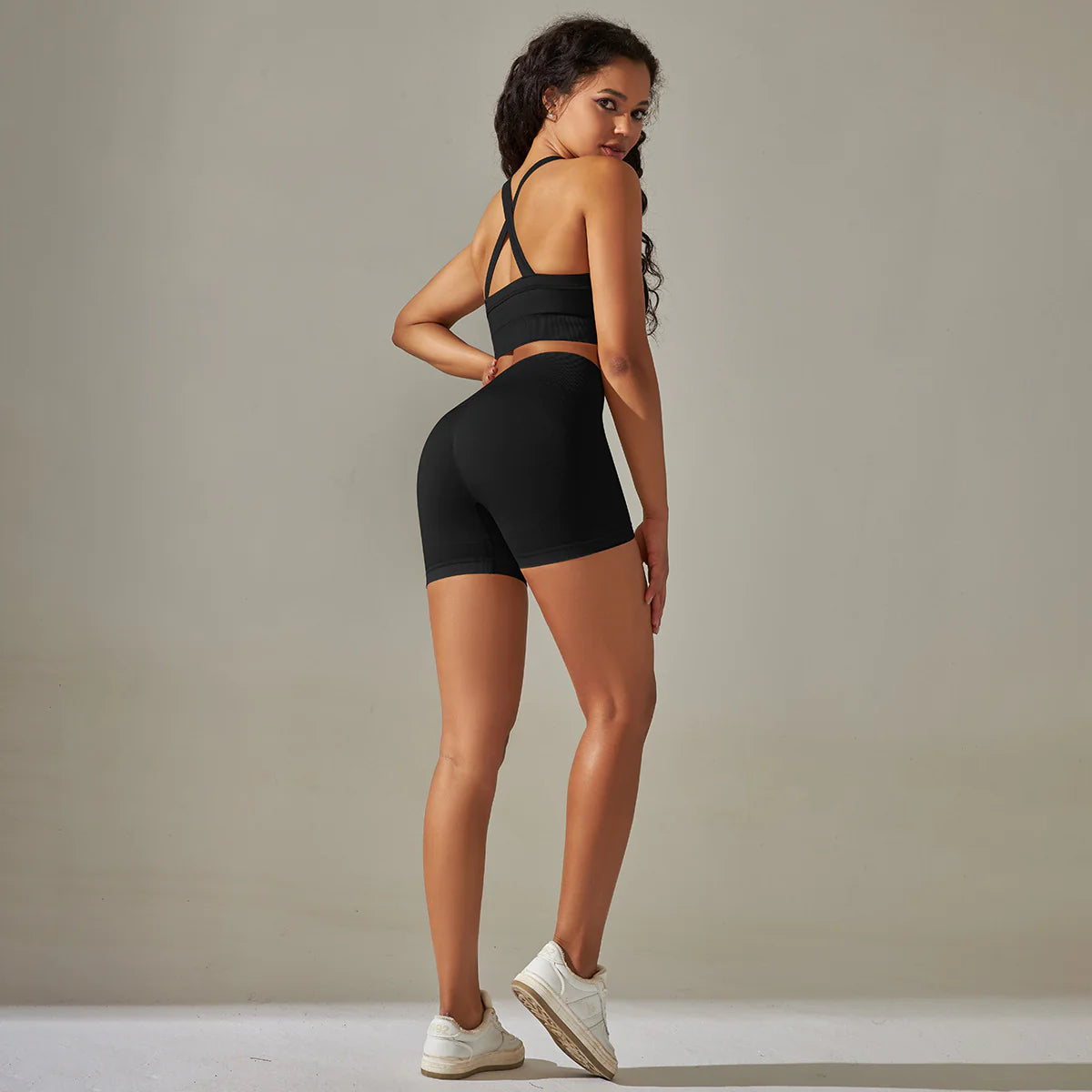 Woman wearing a black sports bra and shorts set with white sneakers on a neutral background