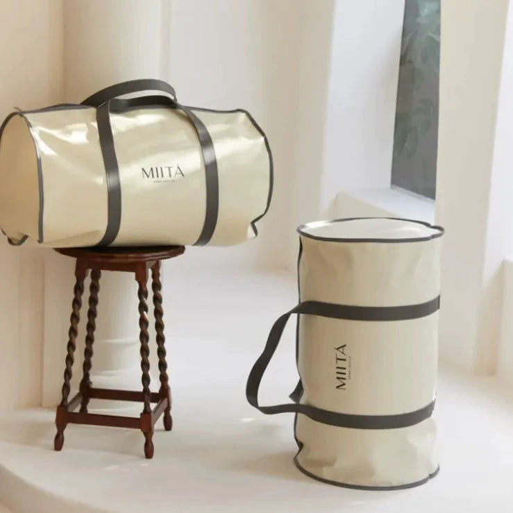 Two beige bags with black straps on a white surface, one on a small wooden table.