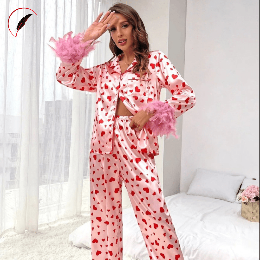 A woman models a two-piece satin pajama set with a red heart pattern on a light pink background, featuring fluffy pink feather-trimmed cuffs. She is standing in a bedroom setting with a bed and white curtains behind her.