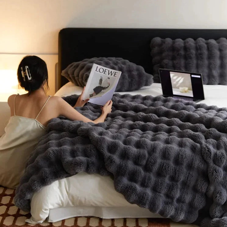 Woman reading a magazine on a bed with a Loewe cover, using a tablet, and covered by a textured gray blanket.